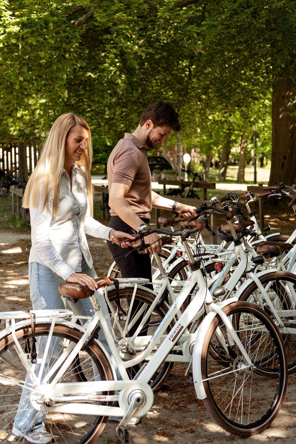 Twee mensen staan ​​bij witte fietsen in een lommerrijk buitengebied waar fietsen worden verhuurd.