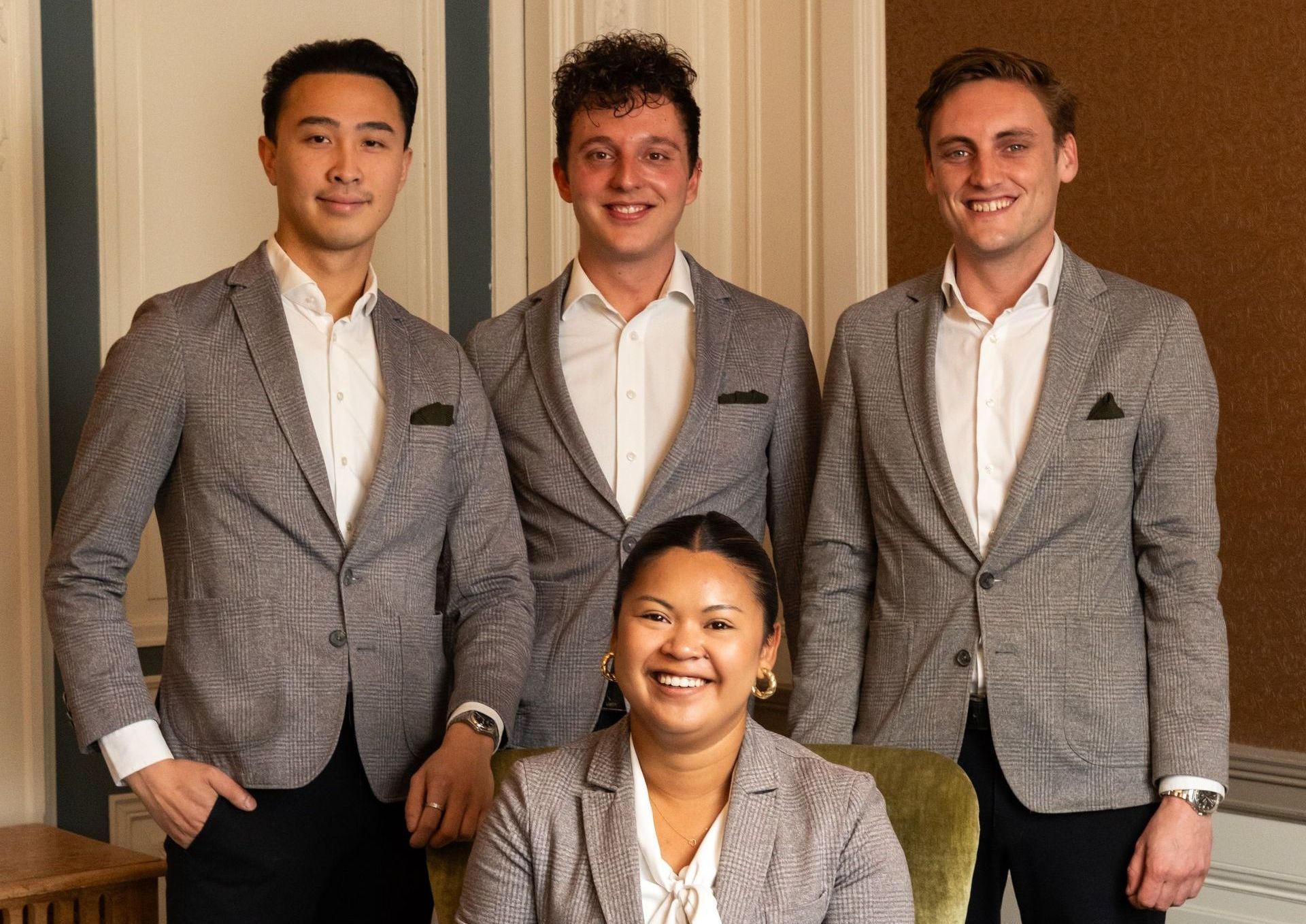 Four people in gray suits posing indoors, three standing behind one seated woman.