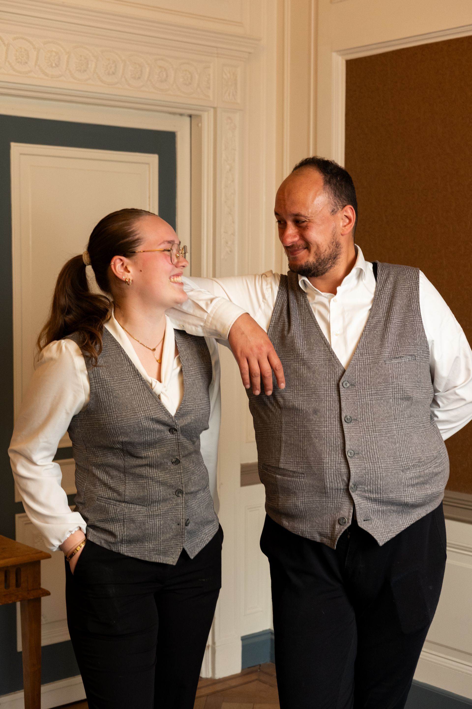 Two people in gray vests smiling and leaning together in a hallway