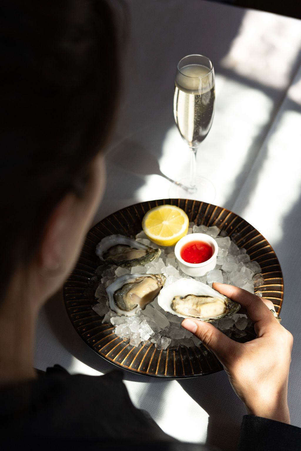 Iemand eet oesters van een bord met citroen, dipsaus en een glas mousserende wijn bij een licht raam.