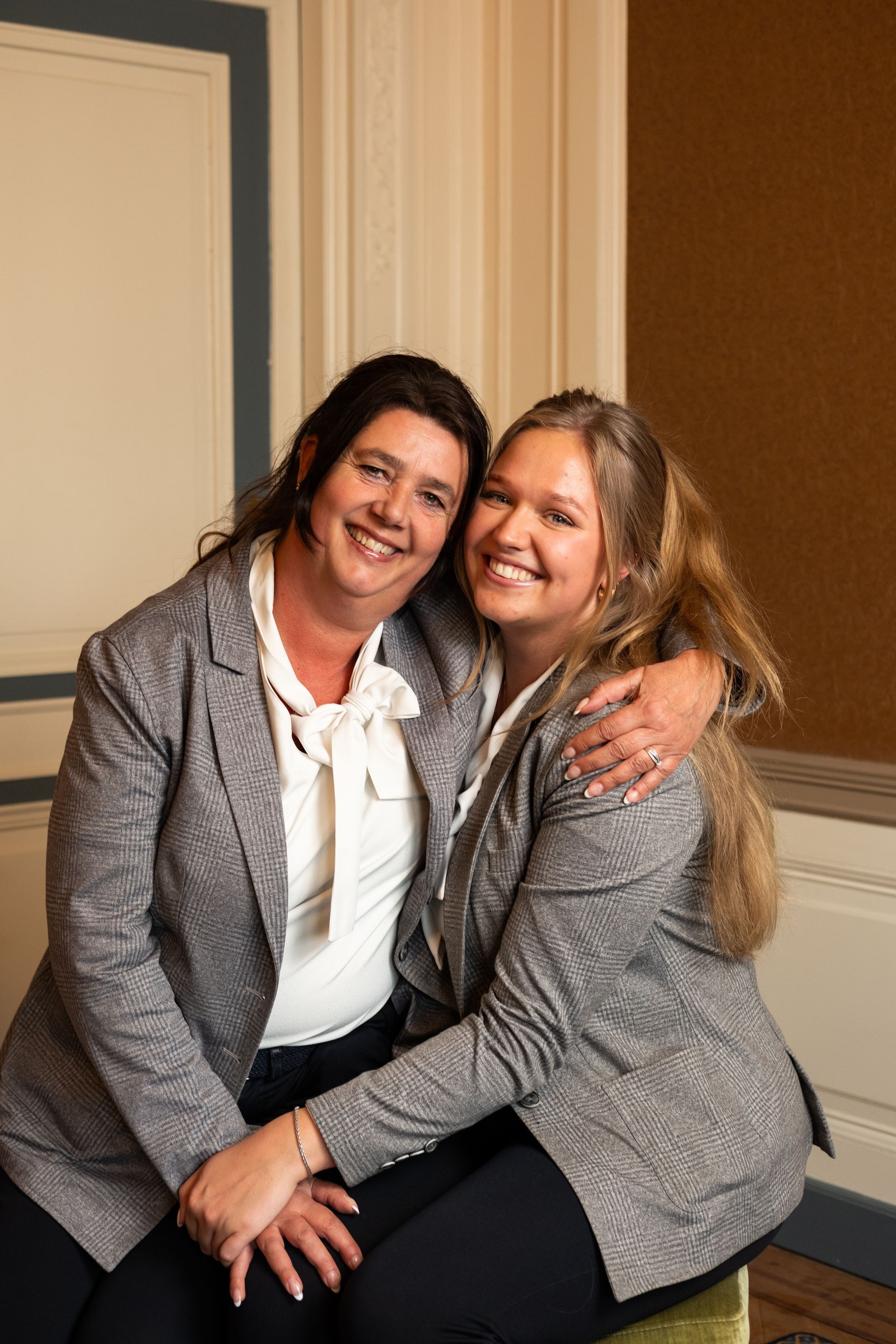 Two smiling women in gray blazers sitting together indoors, one arm around the other.