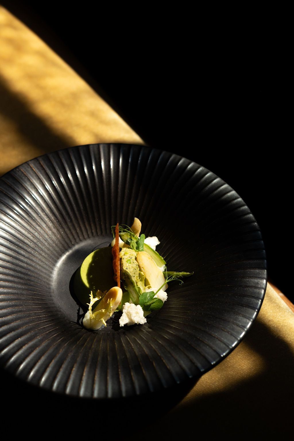 A plated gourmet dish in a black ribbed bowl, lit by sunlight on a dark table.