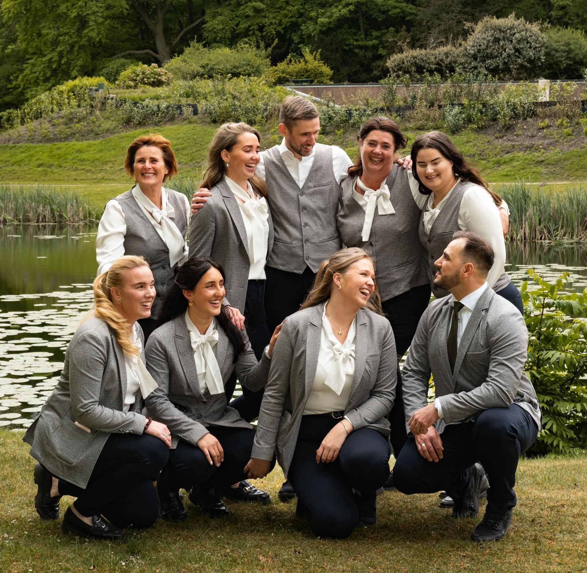 Group of people in gray suits posing outdoors by a pond, smiling in a formal team photo.