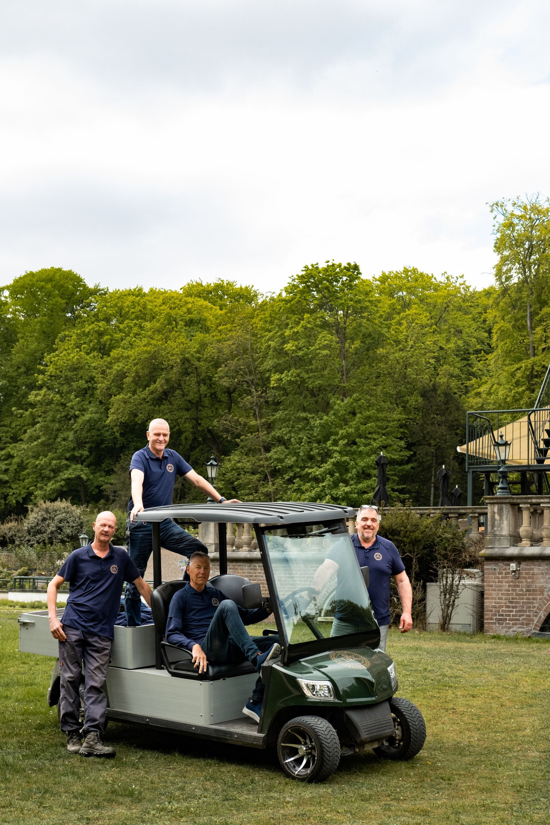 Four people riding a green utility cart on grass beside a hedge and stone wall