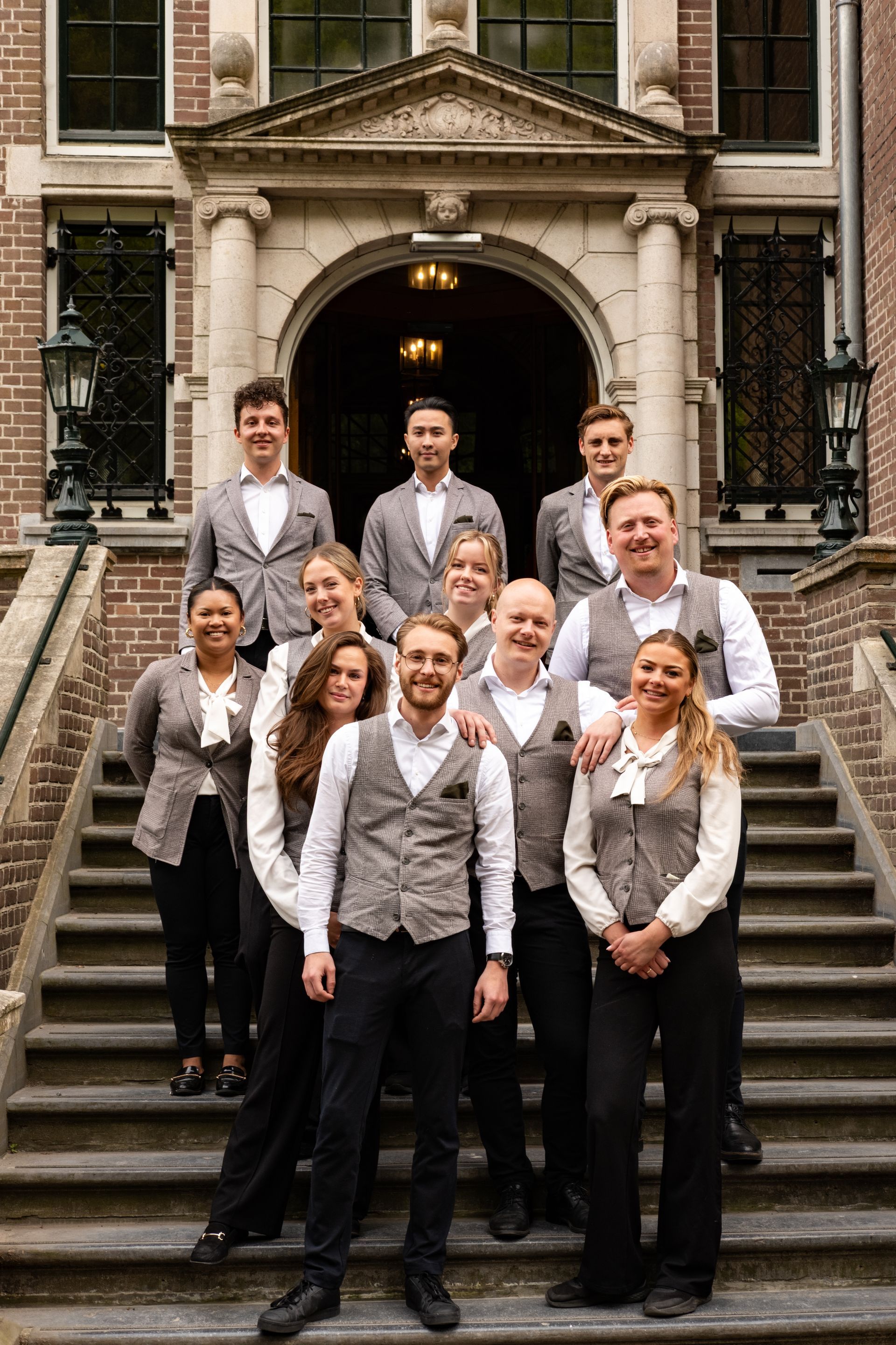 Group of nine people posing on stone steps outside a historic building, wearing coordinated gray and black outfits.