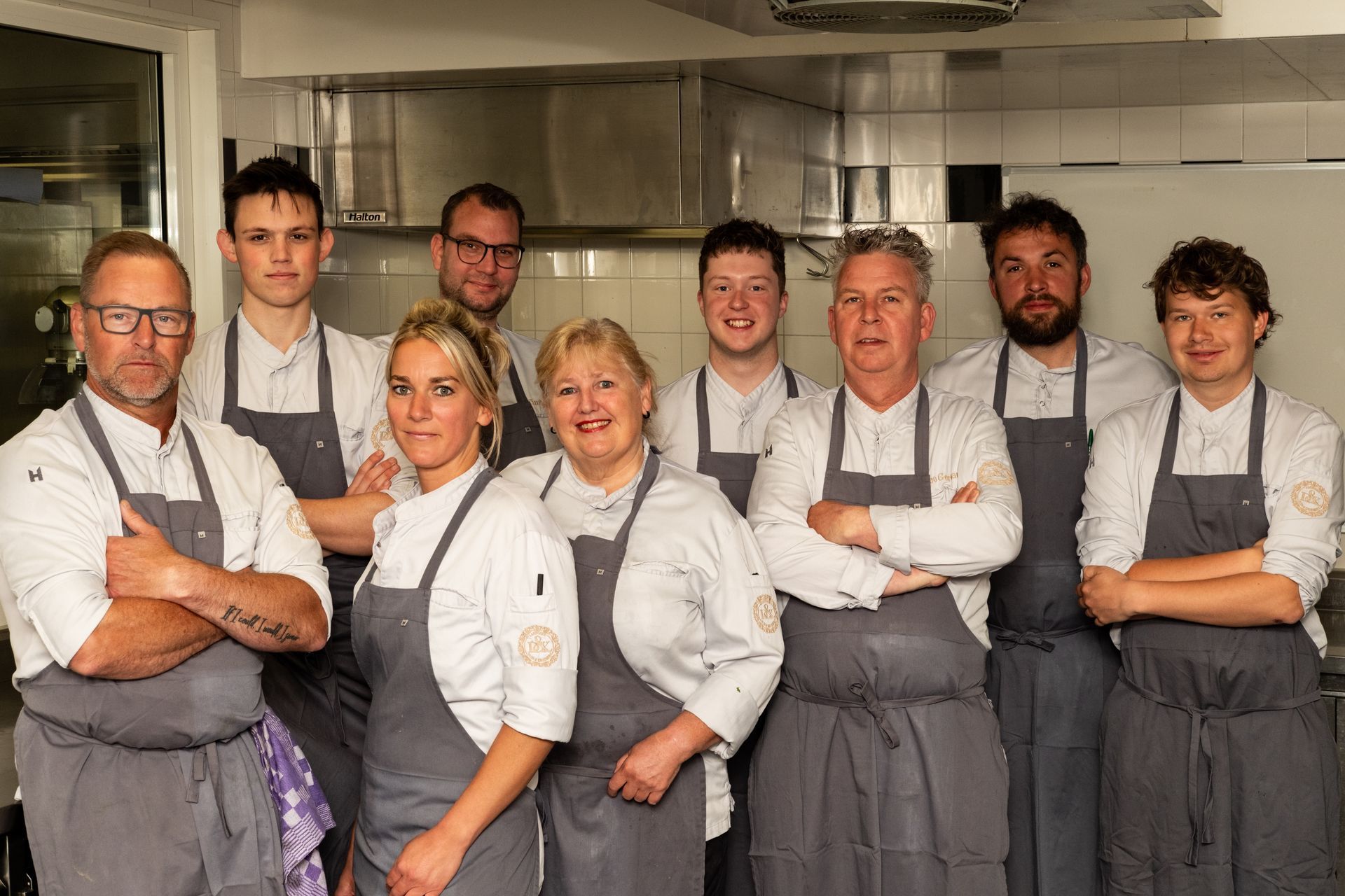 Seven chefs in gray aprons pose together in a commercial kitchen, arms crossed and smiling.