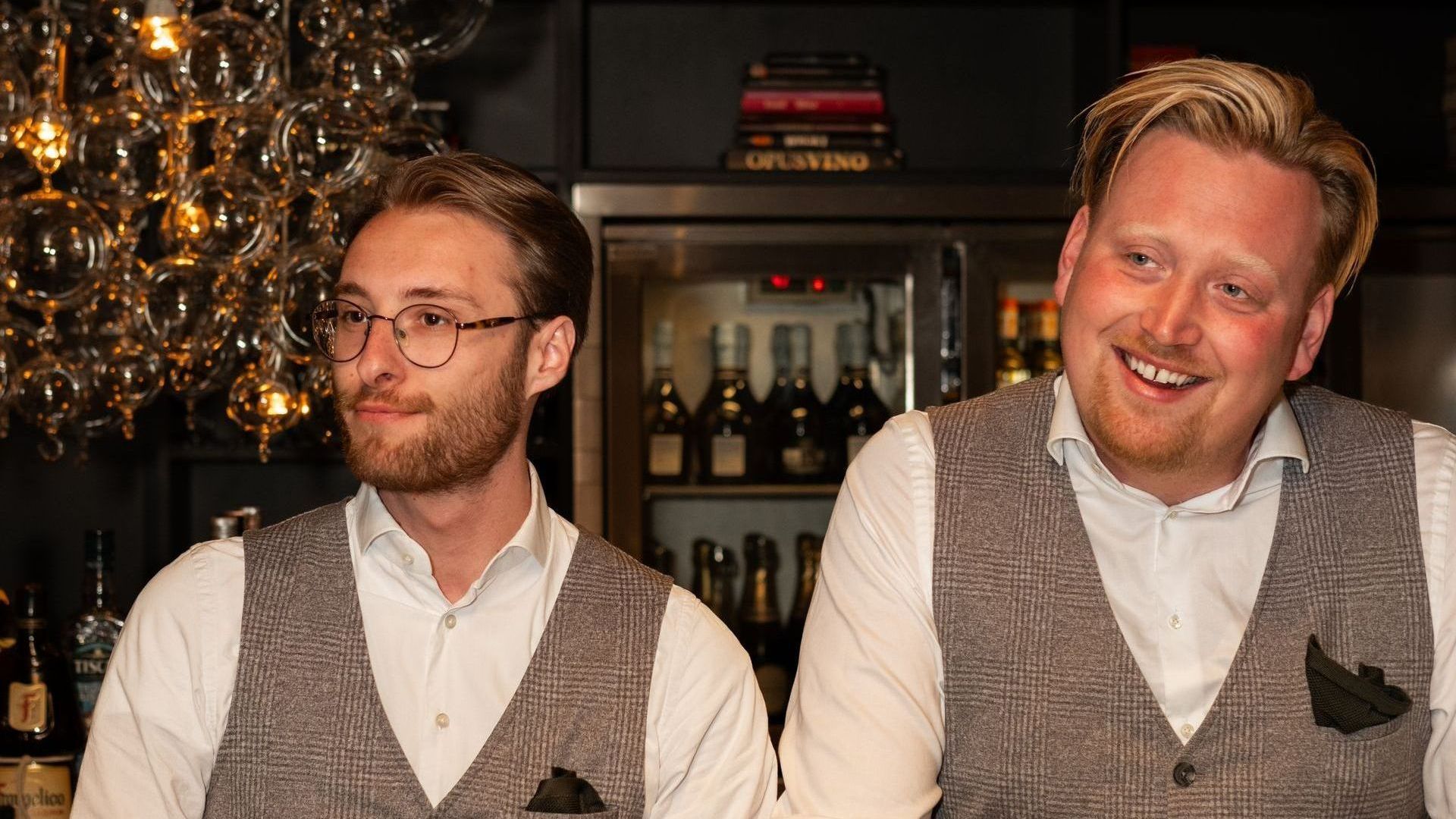 Two men in white shirts and gray vests standing in a warmly lit bar, one smiling at the camera.