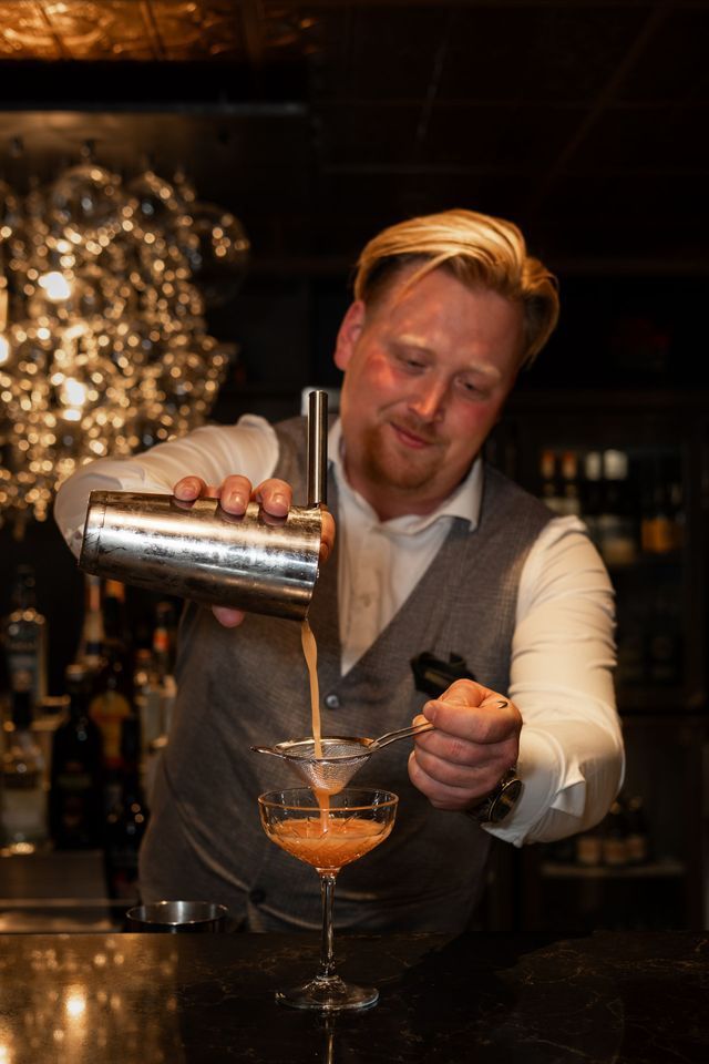 Bartender pouring a cocktail into a coupe glass behind a bar