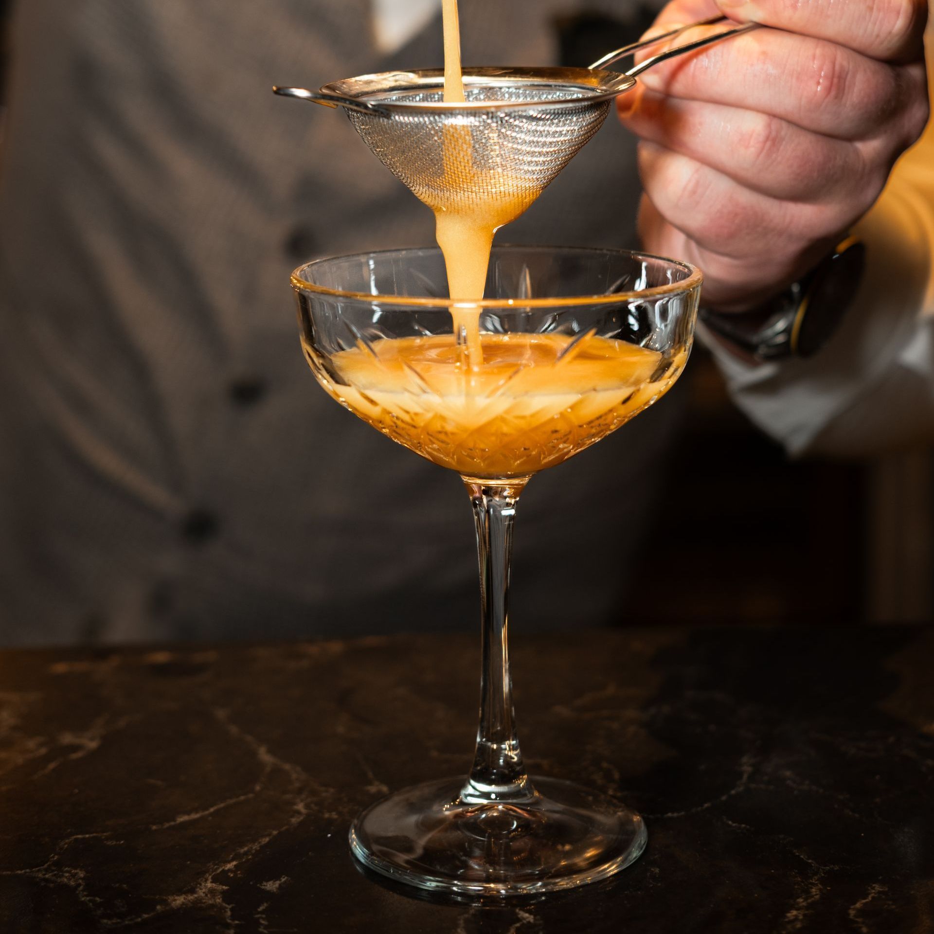 Een cocktail wordt met de hand door een zeef in een coupeglas geschonken aan een bartafel.