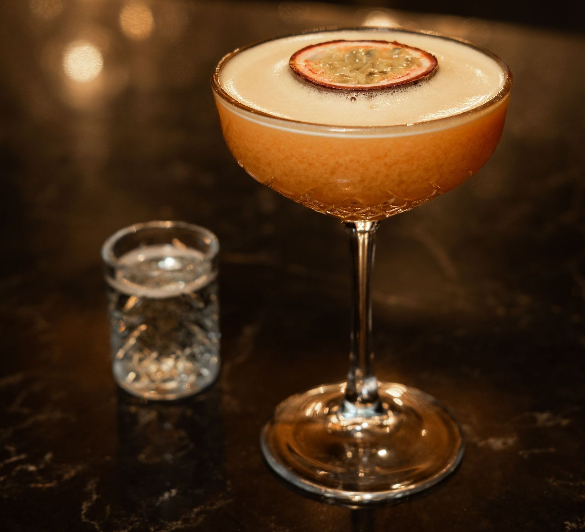 Ein Cocktail in einem Coupé-Glas mit Zitrusgarnitur neben einem Schnapsglas auf einer dunklen Bar.