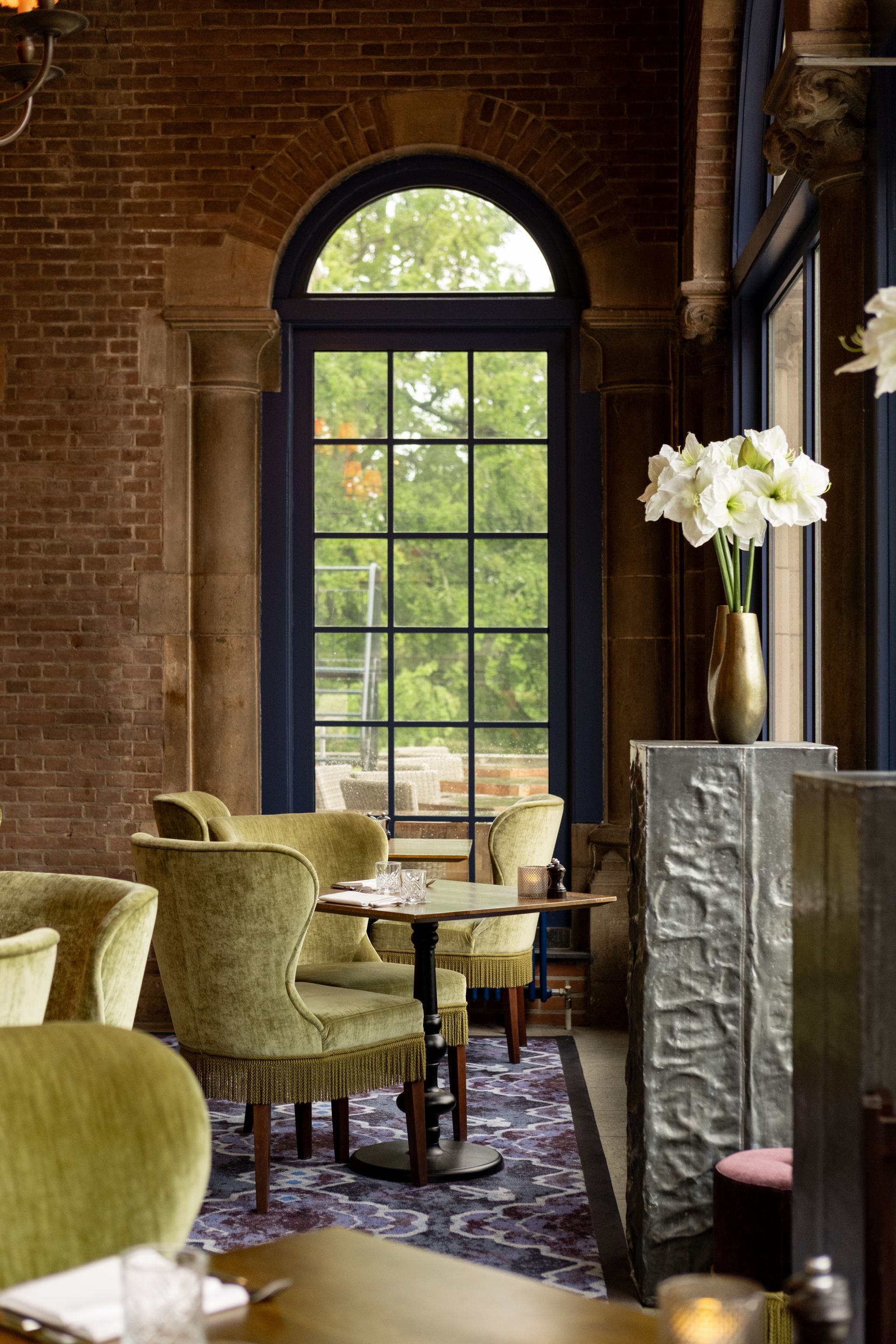 Elegante lounge met groene stoelen, een boogvormig raam, bakstenen muren en witte bloemen bij het raam.