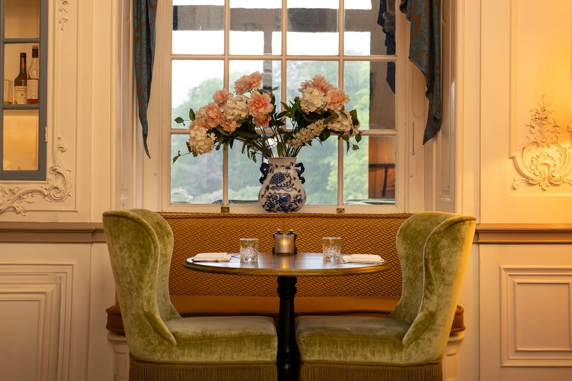 Elegant window-side café table with green velvet chairs, blue vase of flowers, and warm interior lighting