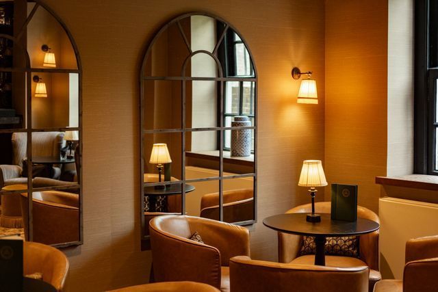 Cozy lounge with tan armchairs, round tables, lamps, and an arched mirror in warm amber light