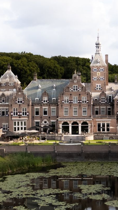 Historisch bakstenen herenhuis aan een vijver, met sierlijke torens en bomen die in het water worden weerspiegeld.