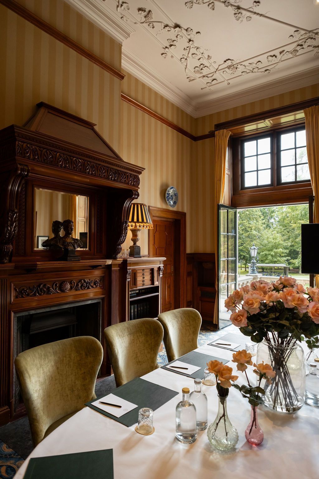 Elegant dining room with ornate wood fireplace, set table, and large window overlooking greenery