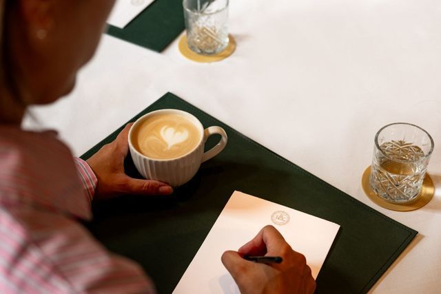 Person writing on a notepad beside a cup of coffee at a table with glasses.