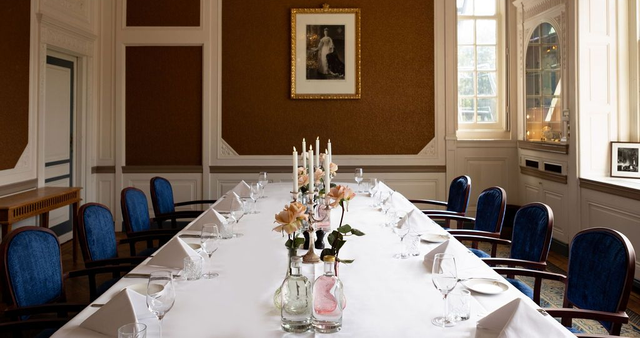 Formele eetkamer met een lange witte tafel, blauwe stoelen en een bloemstuk in het midden bij de hoge ramen.