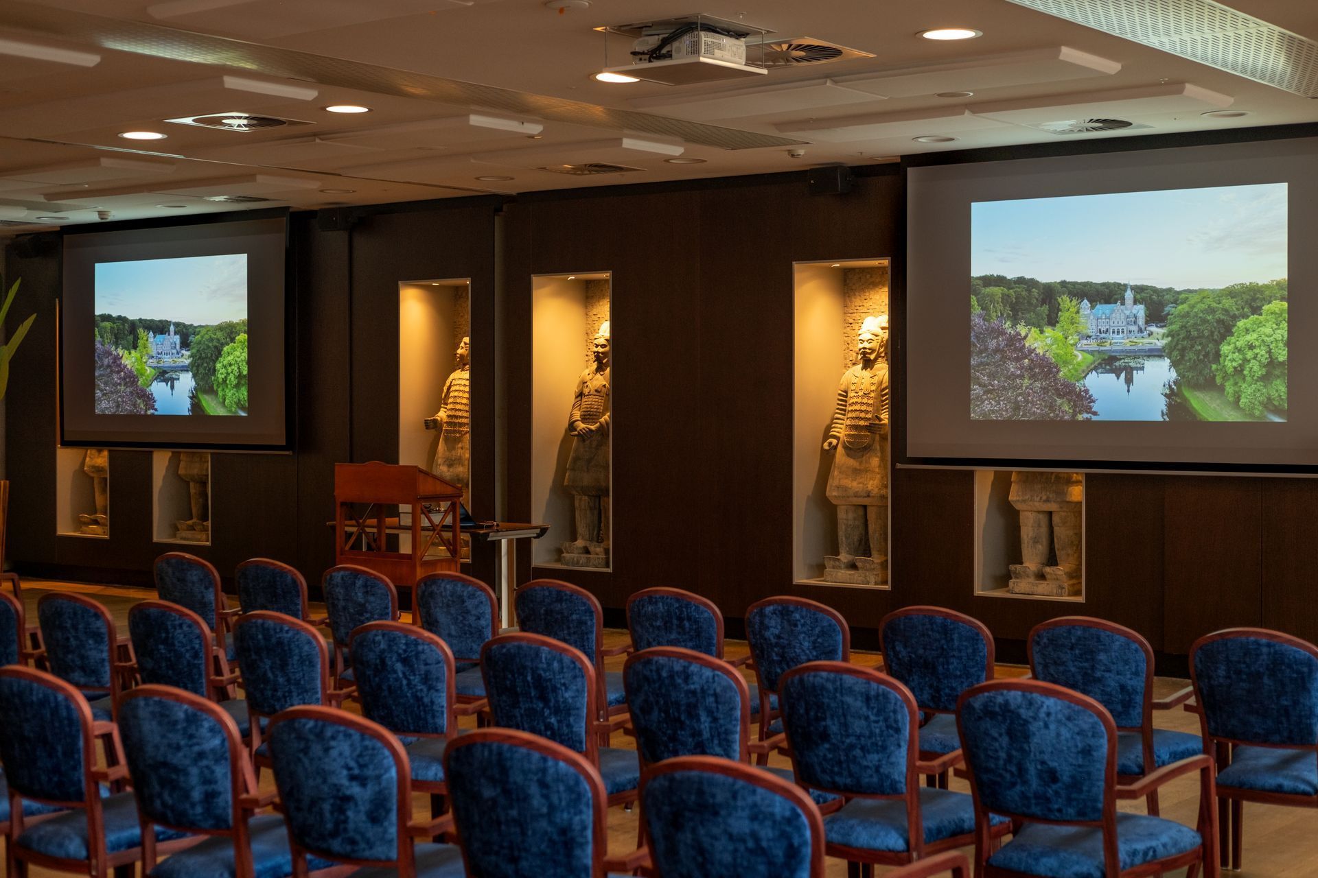 Conference room with rows of blue chairs and two screens displaying a landscape scene.