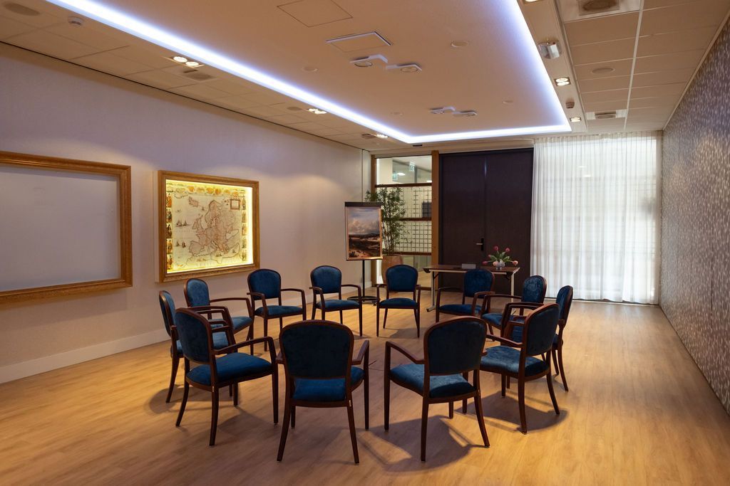 Empty meeting room with round table and blue chairs, wooden floor, and bright window light