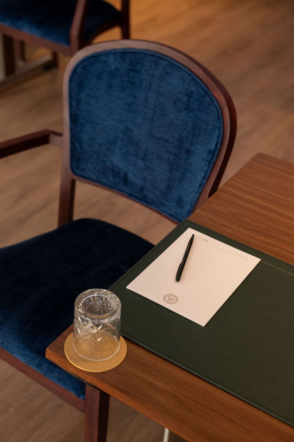 Blue velvet chair beside a wooden table with a green placemat, note card, pen, and glass of water.