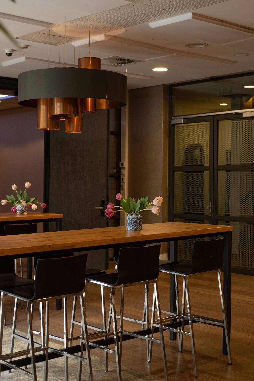Modern office lounge with a long wooden table, black stools, and hanging pendant lights