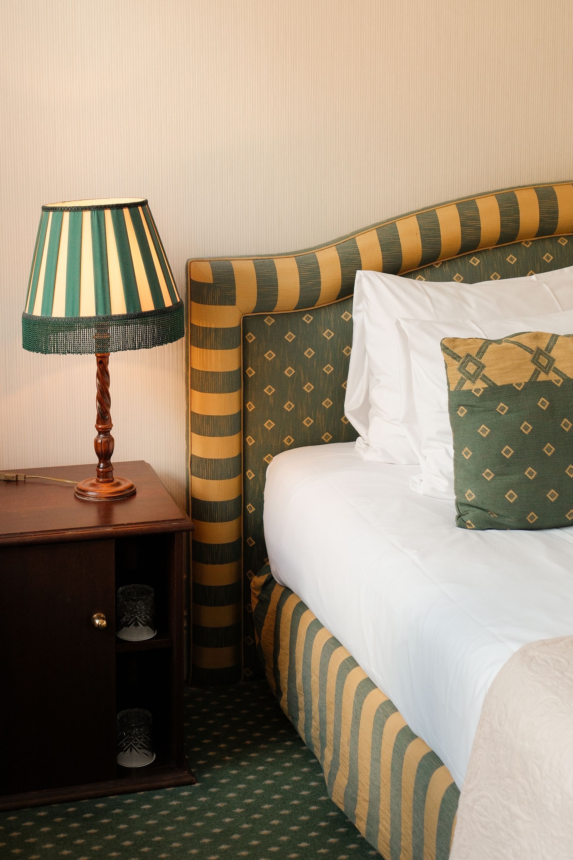Slaapkamerhoek met een gestreept gestoffeerd bed, wit beddengoed, groene kussens en een lamp op een houten nachtkastje.