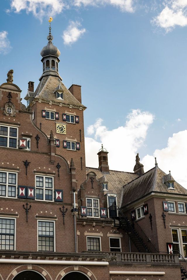 Historisch bakstenen gebouw met sierlijke toren en kleine ramen tegen een blauwe hemel met wolken.