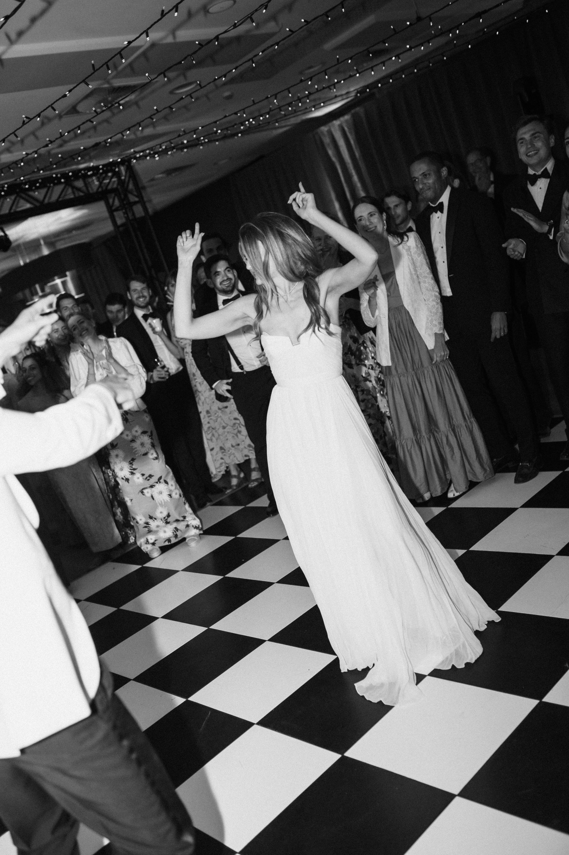 Mensen dansen op een zwart-wit geblokte vloer tijdens een feest, een van hen in een lange witte jurk.