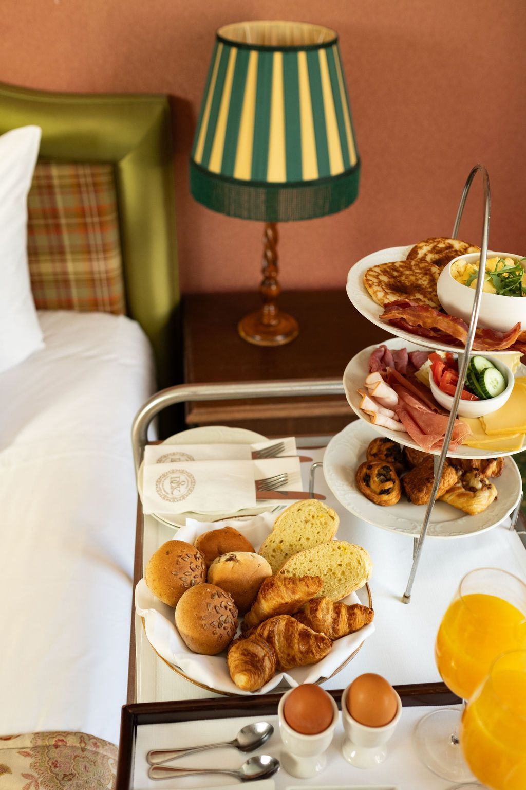 Breakfast tray with pastries, eggs, and orange juice beside a bed and striped lamp