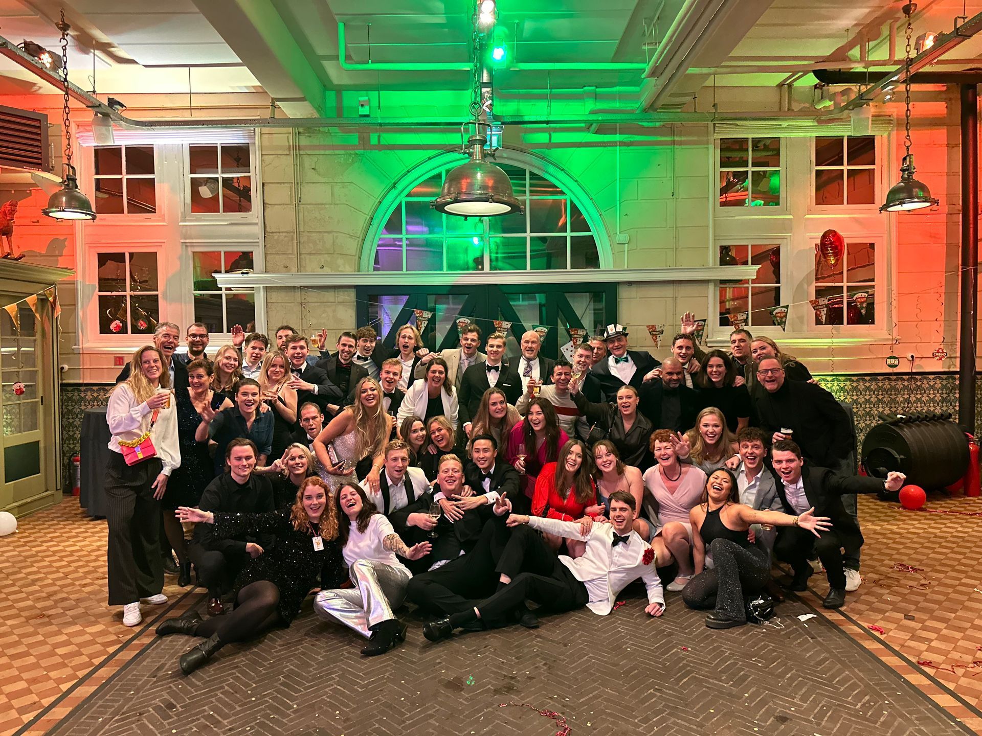Large group posing indoors under green and red lights, smiling in a decorated hall
