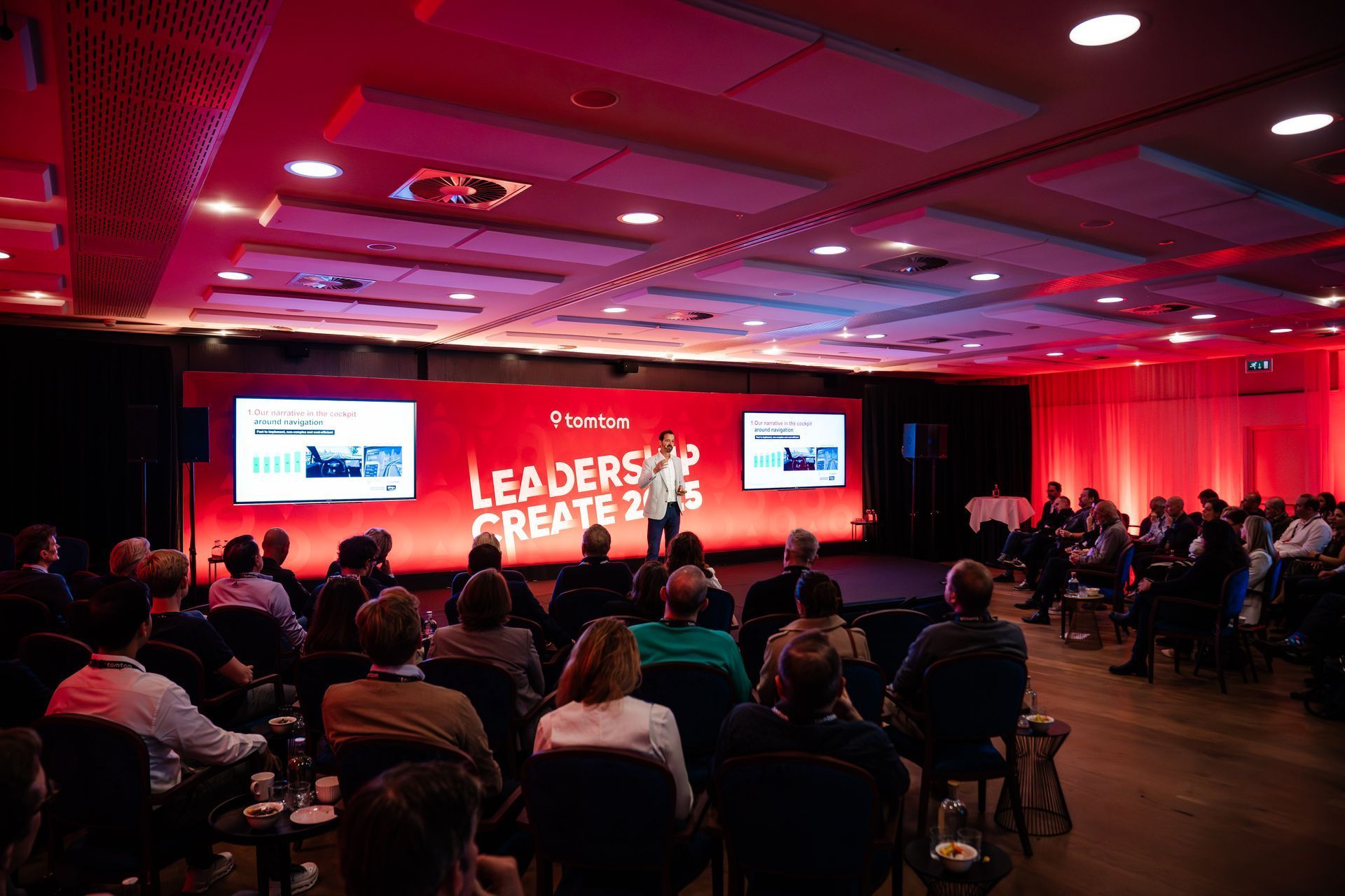 Conference audience watching a red-lit leadership event on a stage with screens and speakers