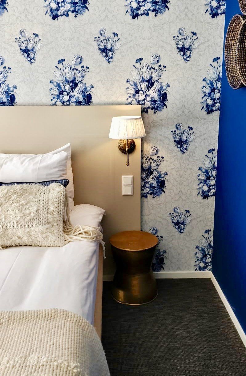 Een kleine slaapkamerhoek met blauw bloemenbehang, een wit bed, een gouden lamp en een houten bijzettafel.