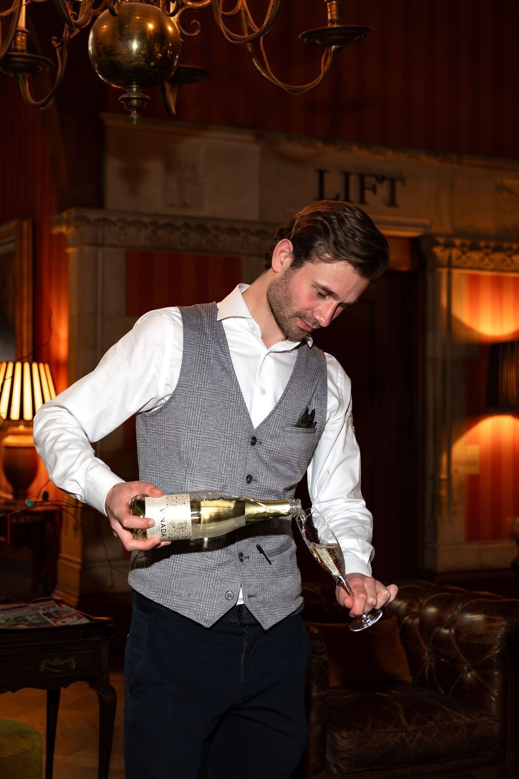 Man in vest pouring champagne at a formal event in a warmly lit room