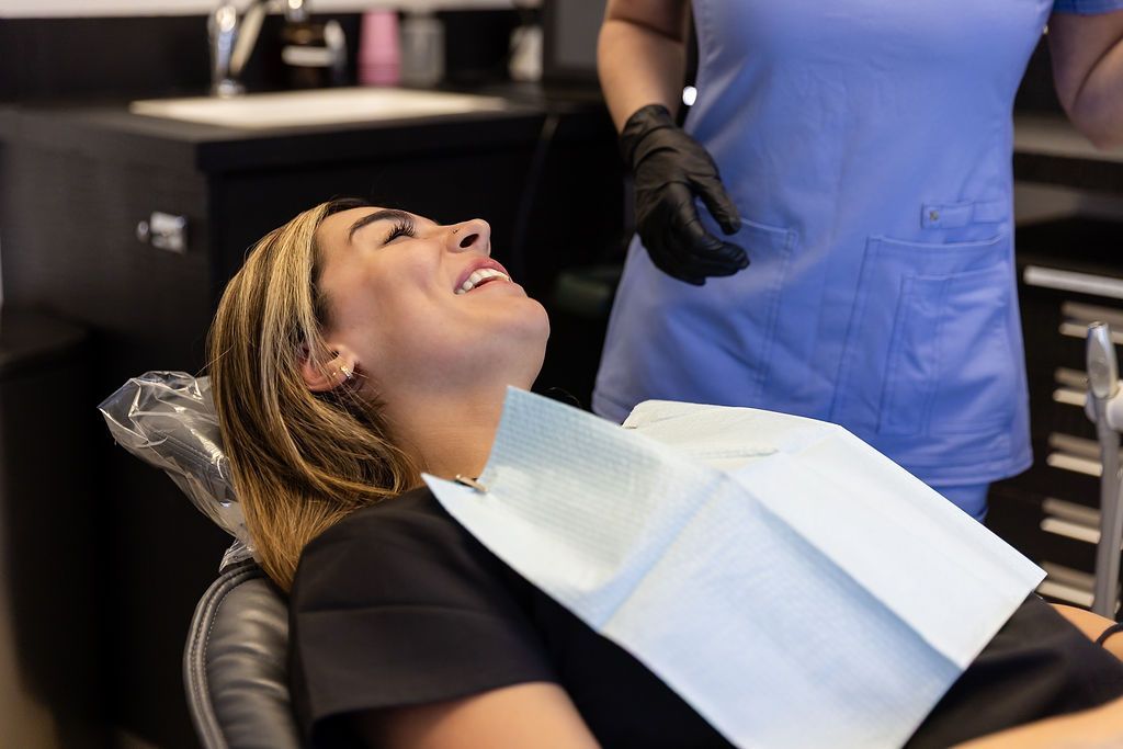 Patient reclined in a dental chair while a dentist works nearby, with a blue bib and gloves visible.
