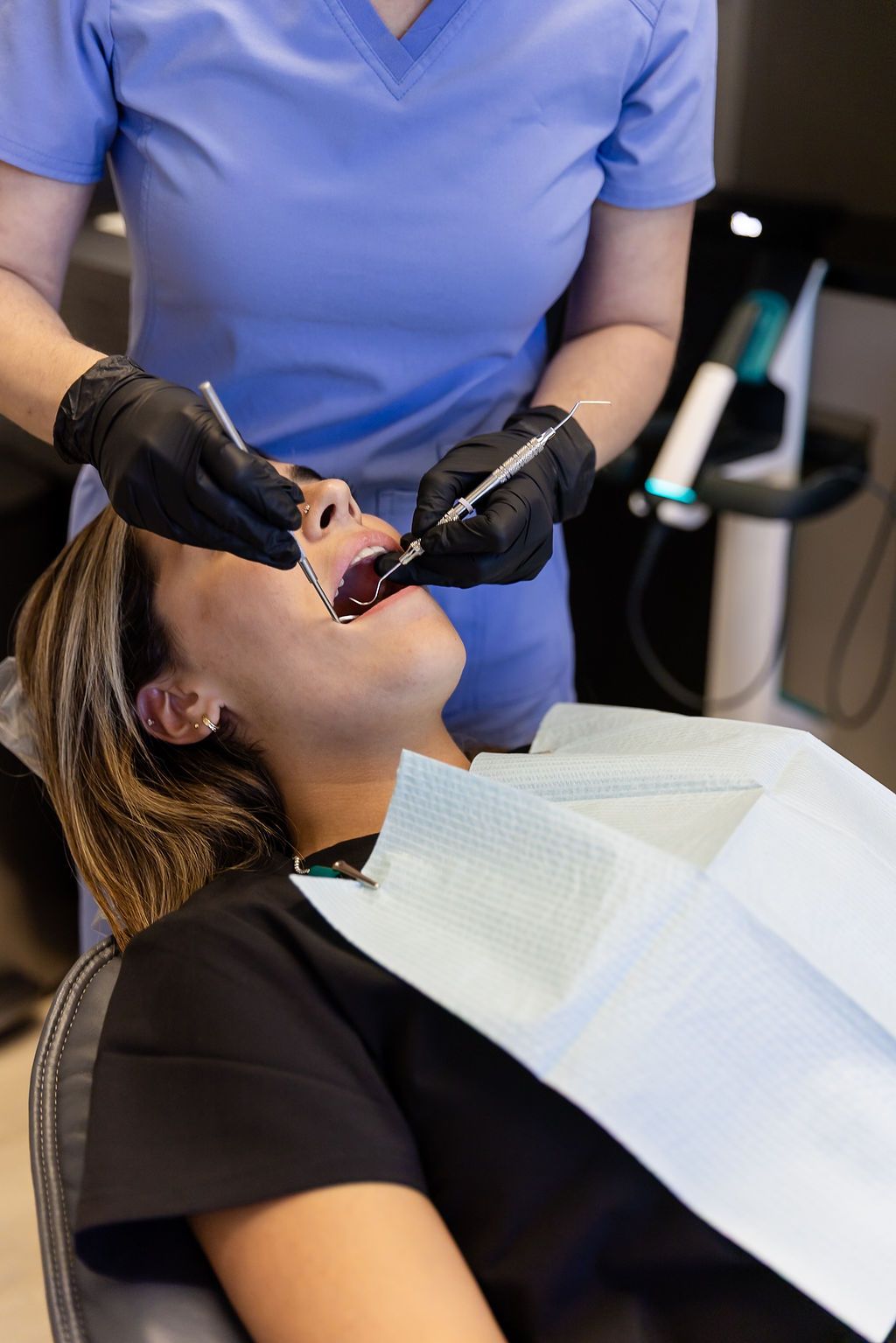 Dentist wearing black gloves examines a patient in a dental chair with a blue bib.