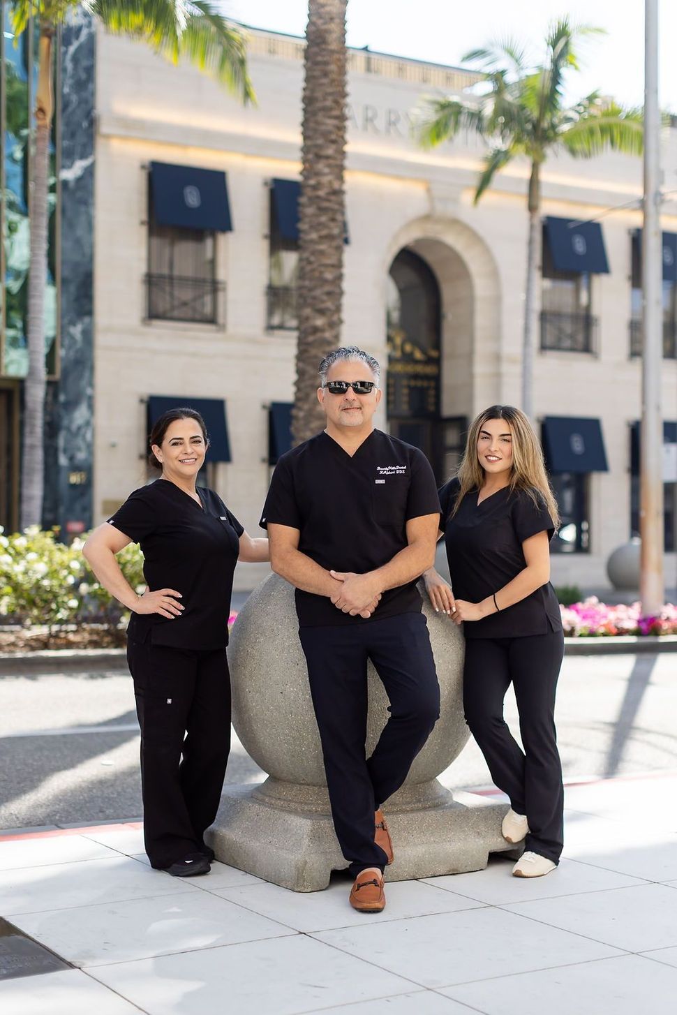 Three people in black outfits posing outdoors by a palm tree and building entrance.