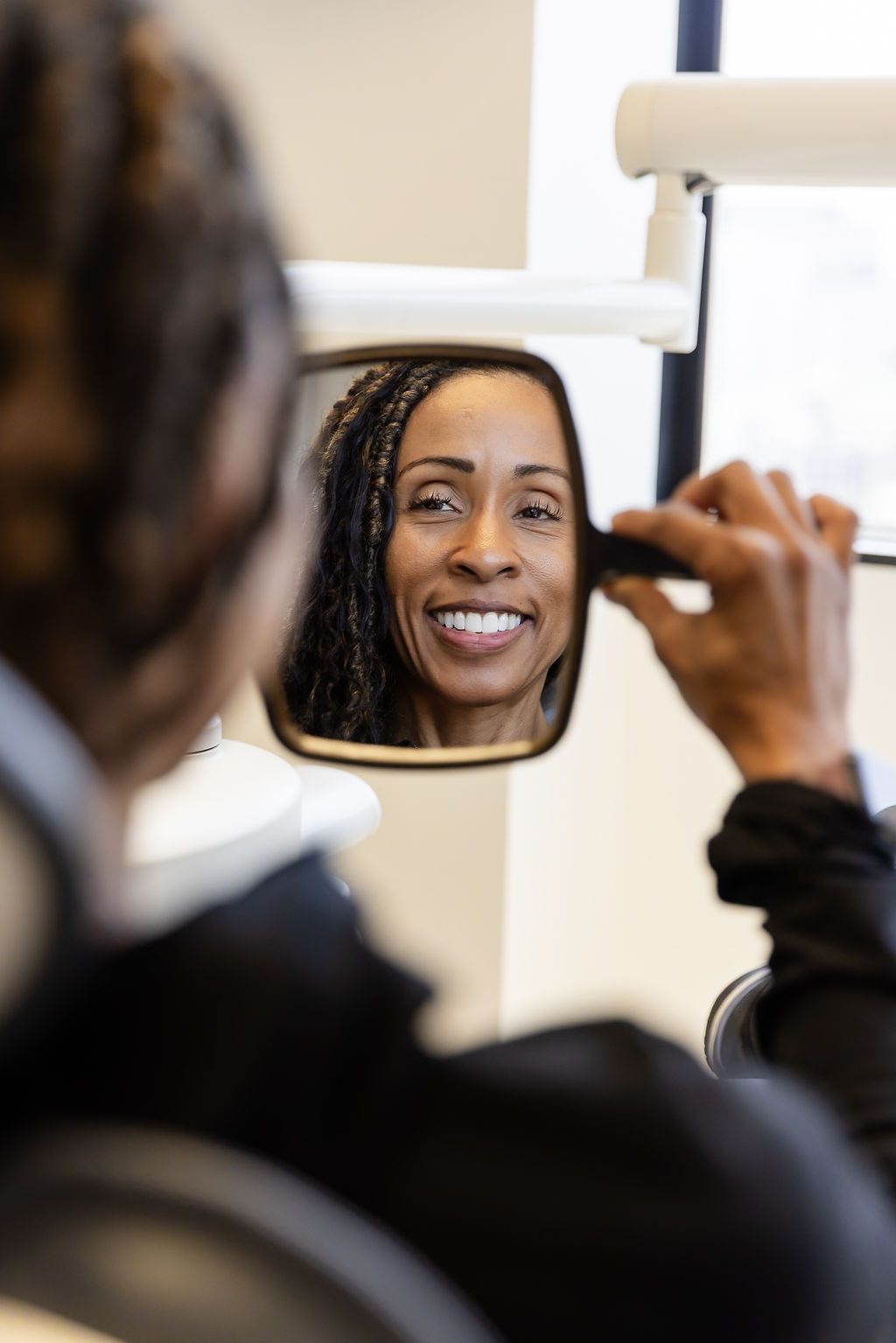 Person smiling into a handheld mirror while adjusting their face, with a blurred reflection in the foreground