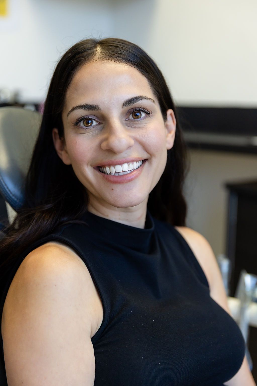 Smiling woman in a black sleeveless top, seated indoors with a blurred background.