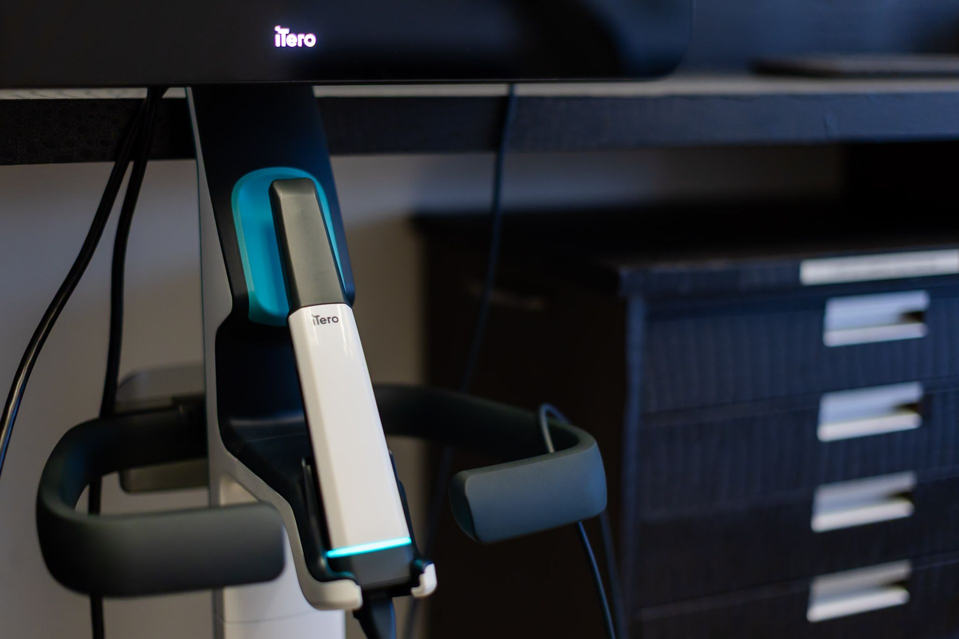 A teal-and-white dental equipment mounted on a black stand beneath a monitor
