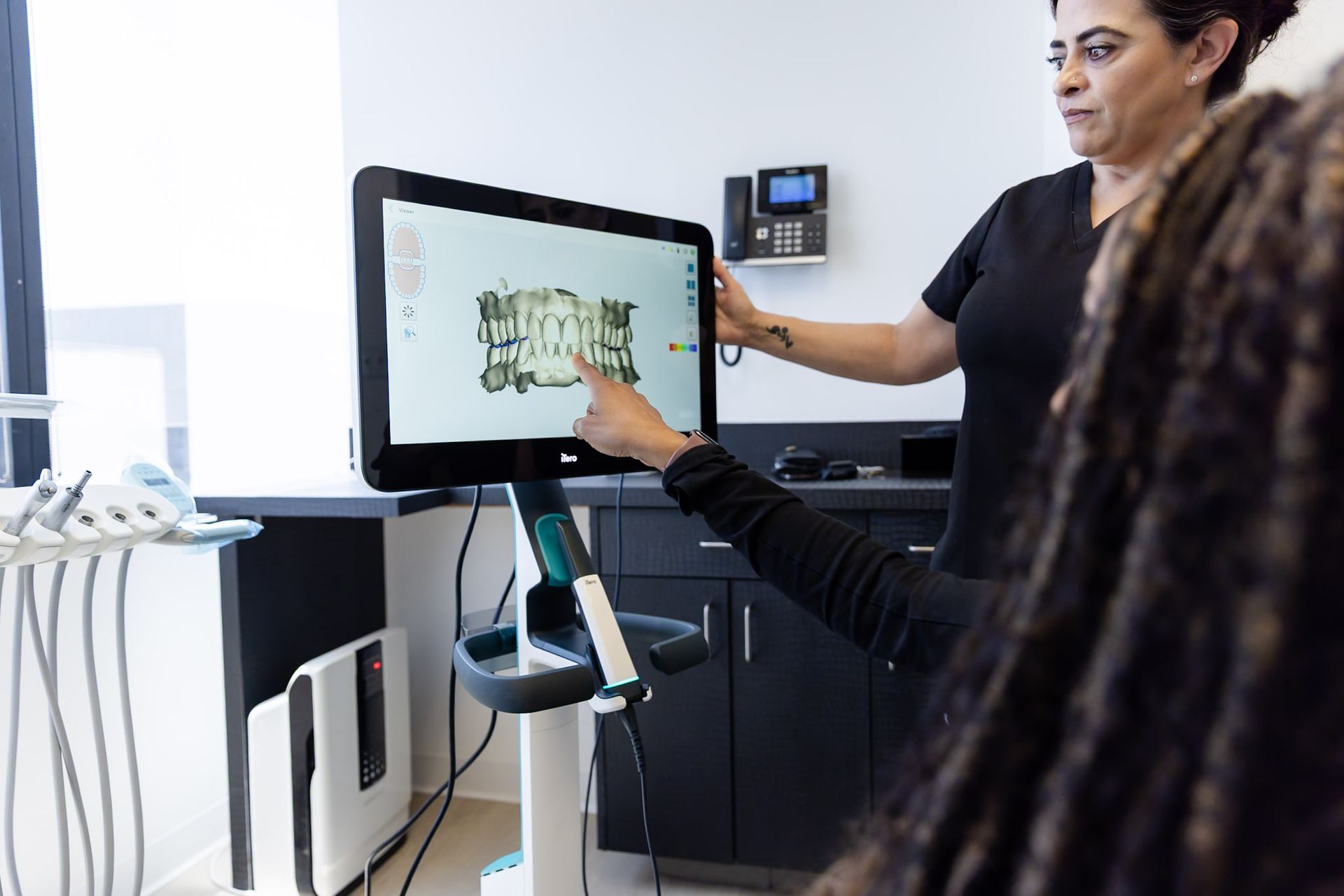Person using a touchscreen kiosk while another person points at the screen in a bright office setting