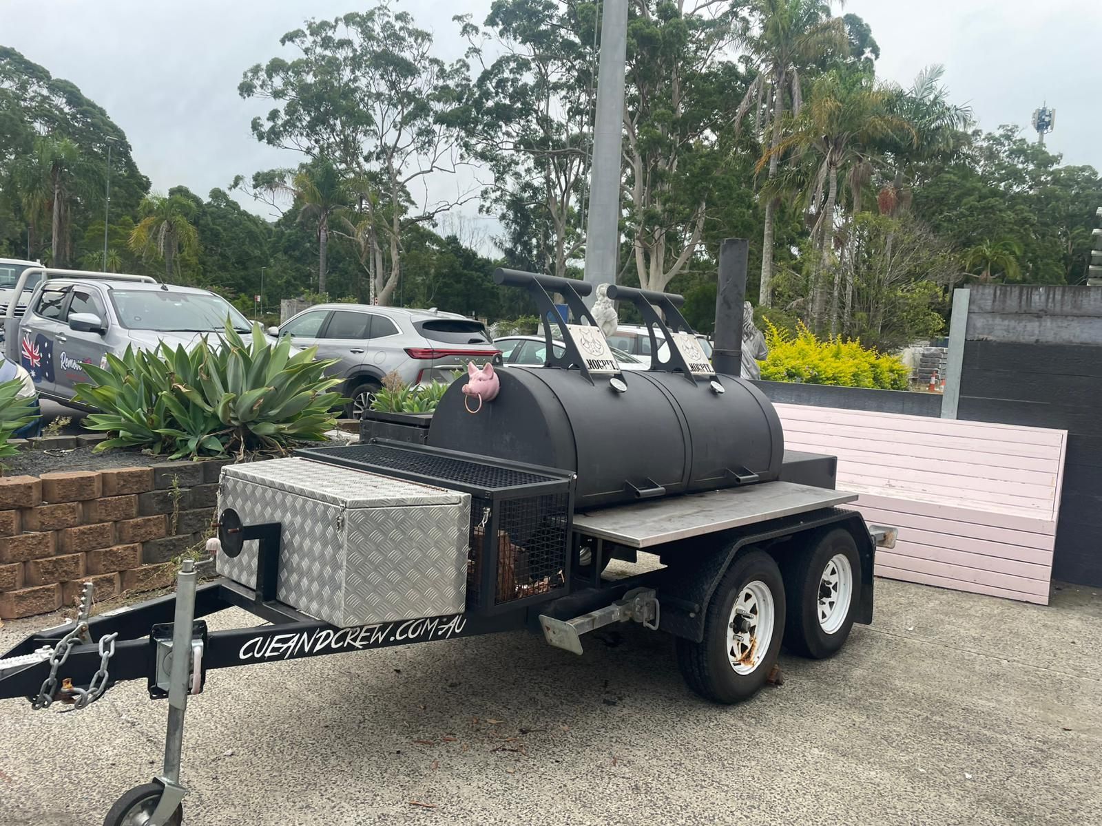 A large, black cylindrical smoker trailer with a silver diamond-plate storage box parked in an outdoor lot — Rustique on the Coast In Ourimbah, NSW