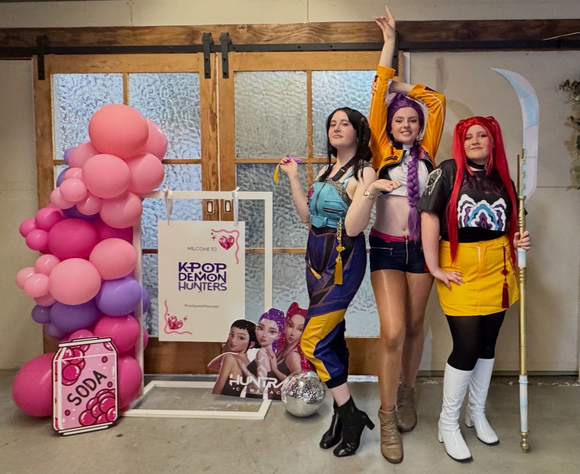 Three people in costumes pose by a sign and balloons, posing as a Kpop group with props.