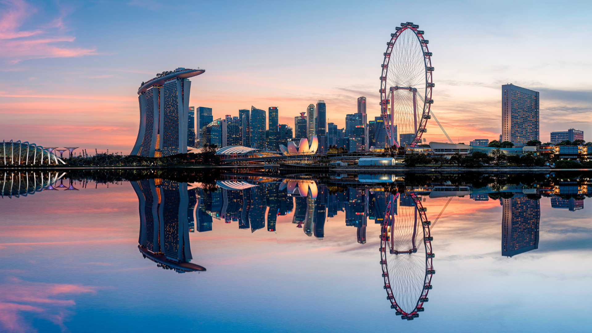 Skyline with a Ferris wheel reflected in calm water at sunset.