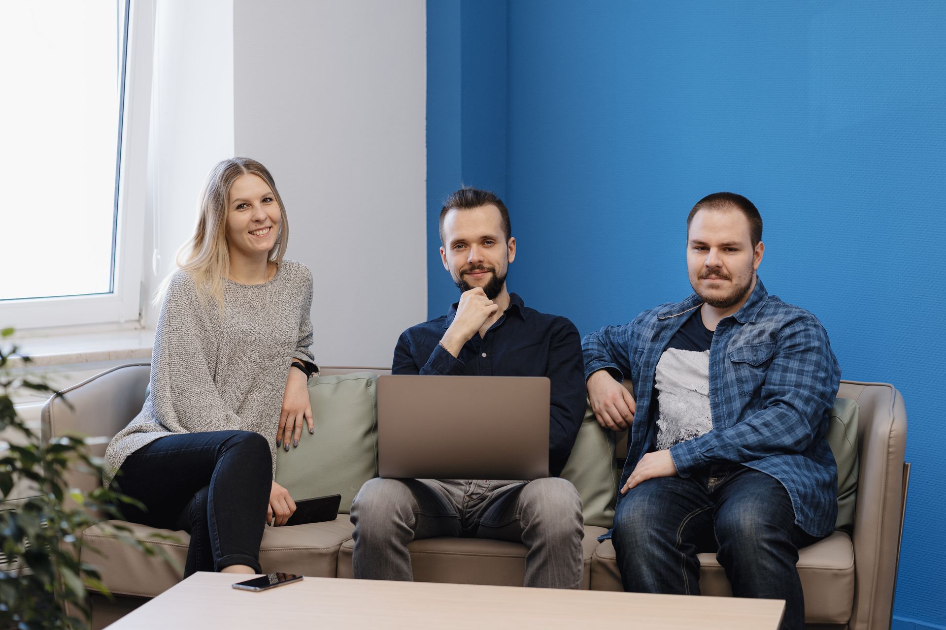 Three people sitting on a couch; woman with blonde hair, two men with laptops, in a blue office.