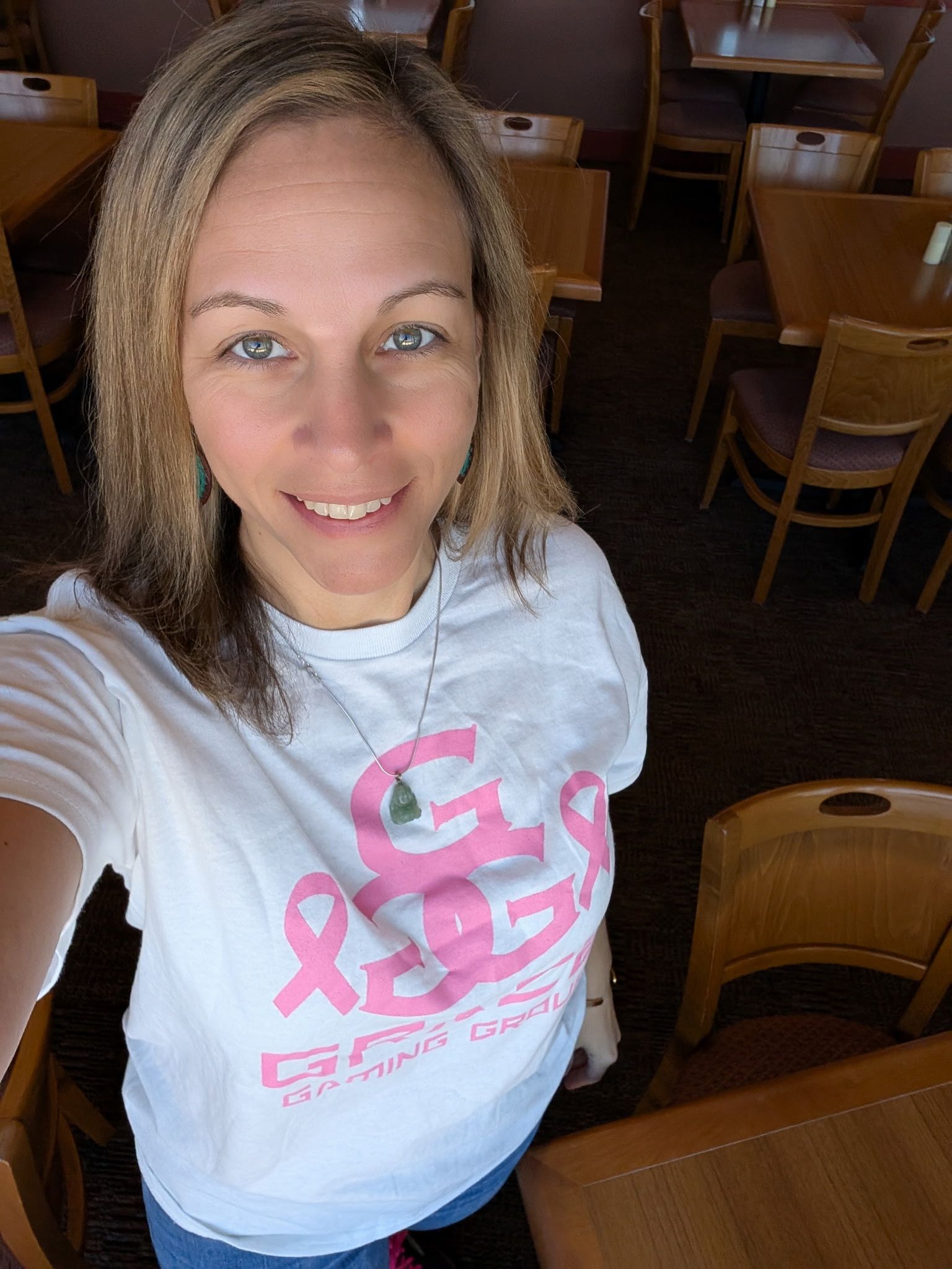 Woman taking a selfie in a restaurant, wearing a white shirt with pink lettering and smiling.