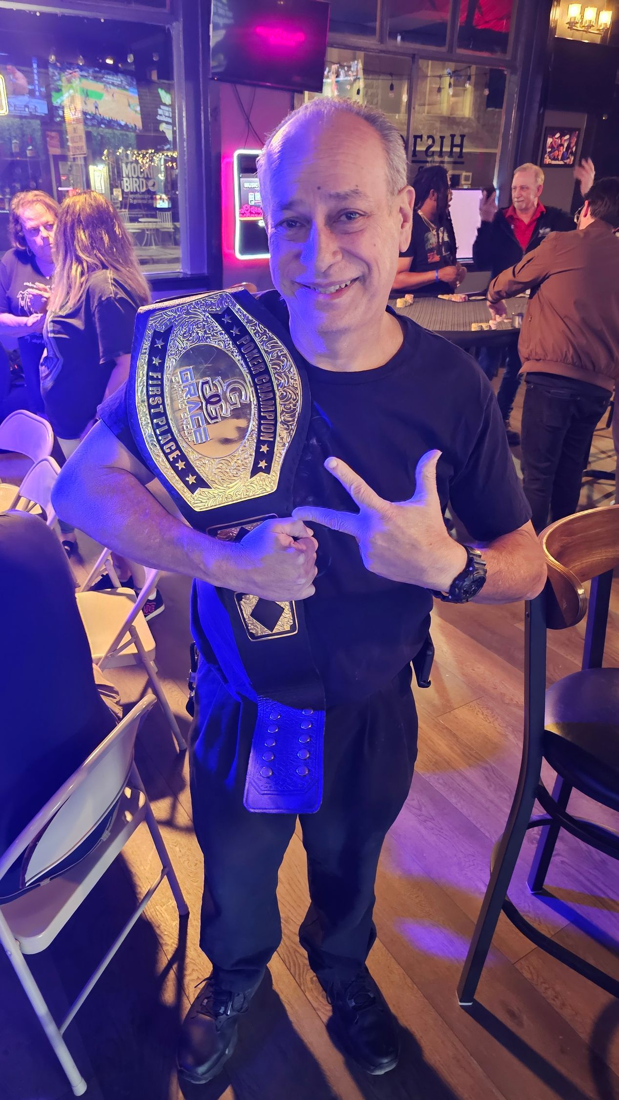 Smiling person holding a championship belt in a dimly lit bar, with chairs and patrons in the background