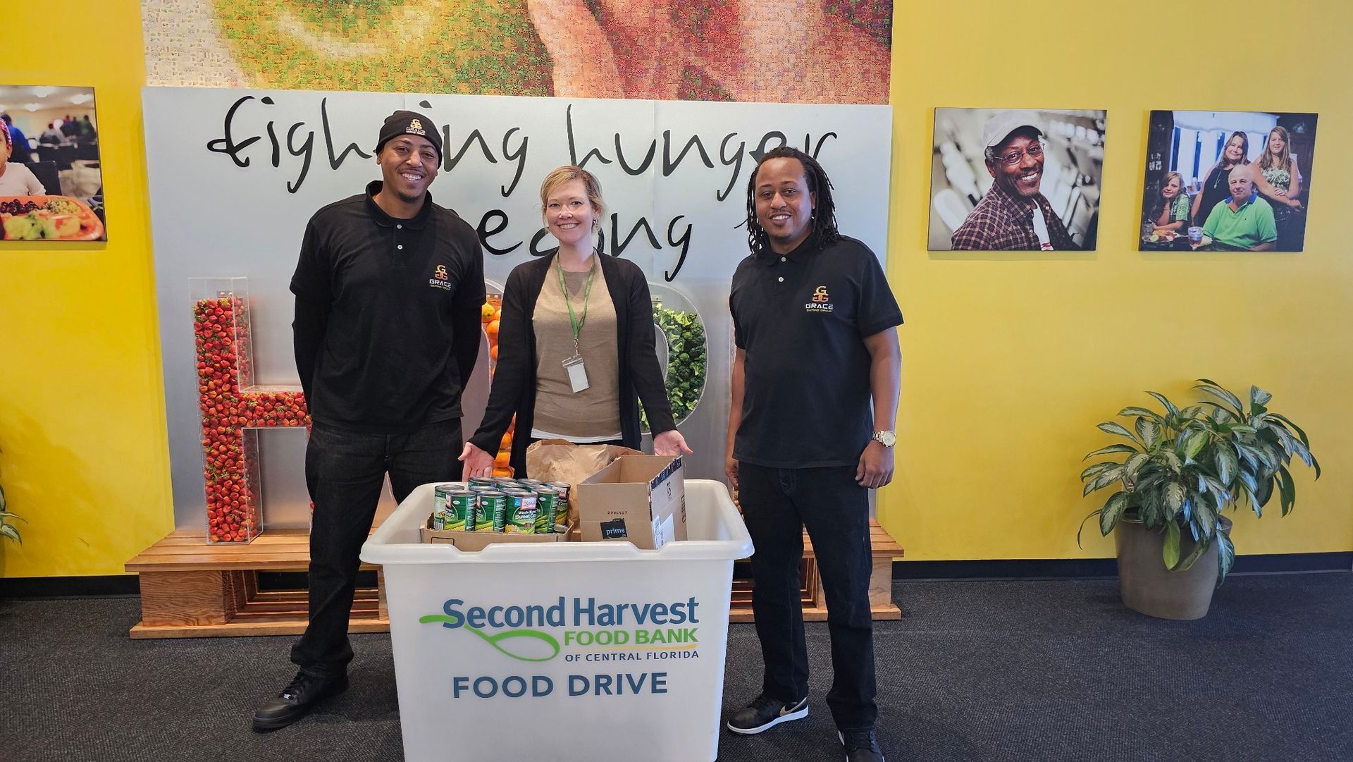 Three people stand behind a table labeled “Second Harvest Food Drive” in a lobby with yellow walls and artwork.