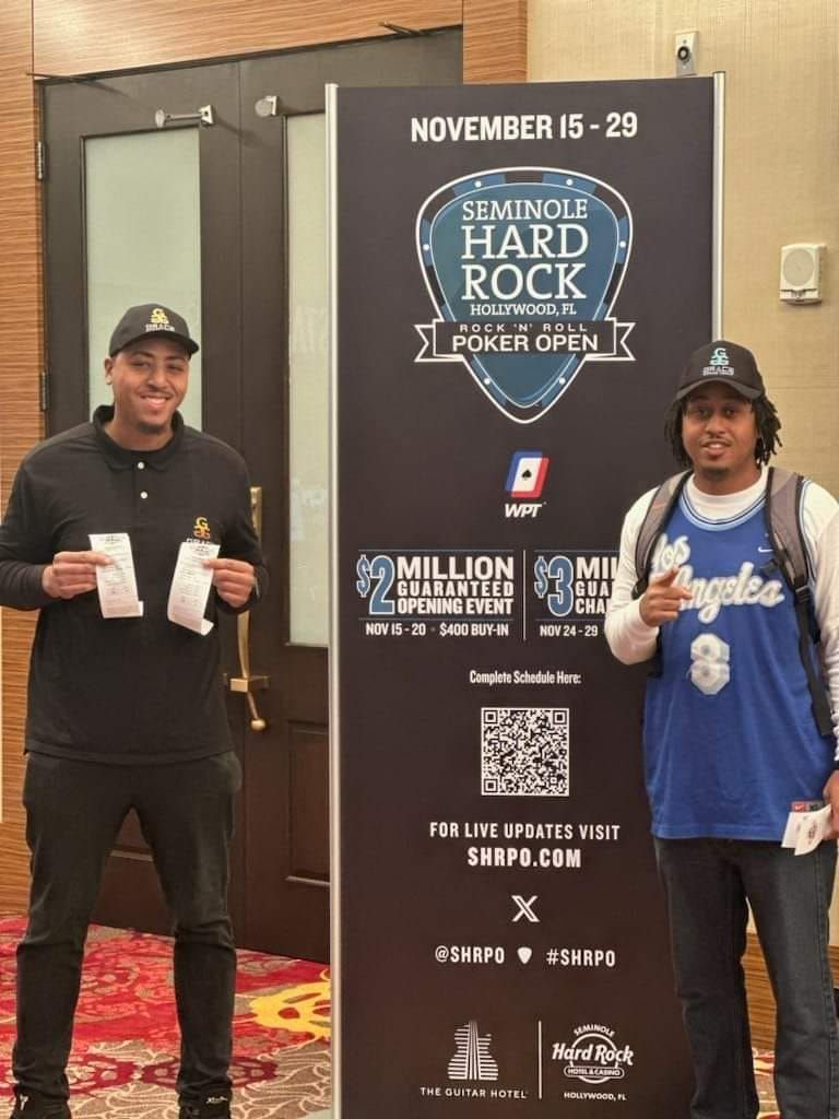 Two men posing with drinks beside a Seminole Hard Rock poker event sign.