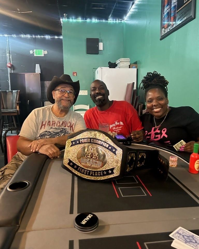 Three people smiling at a pool table with a championship belt laid across it in a game room.