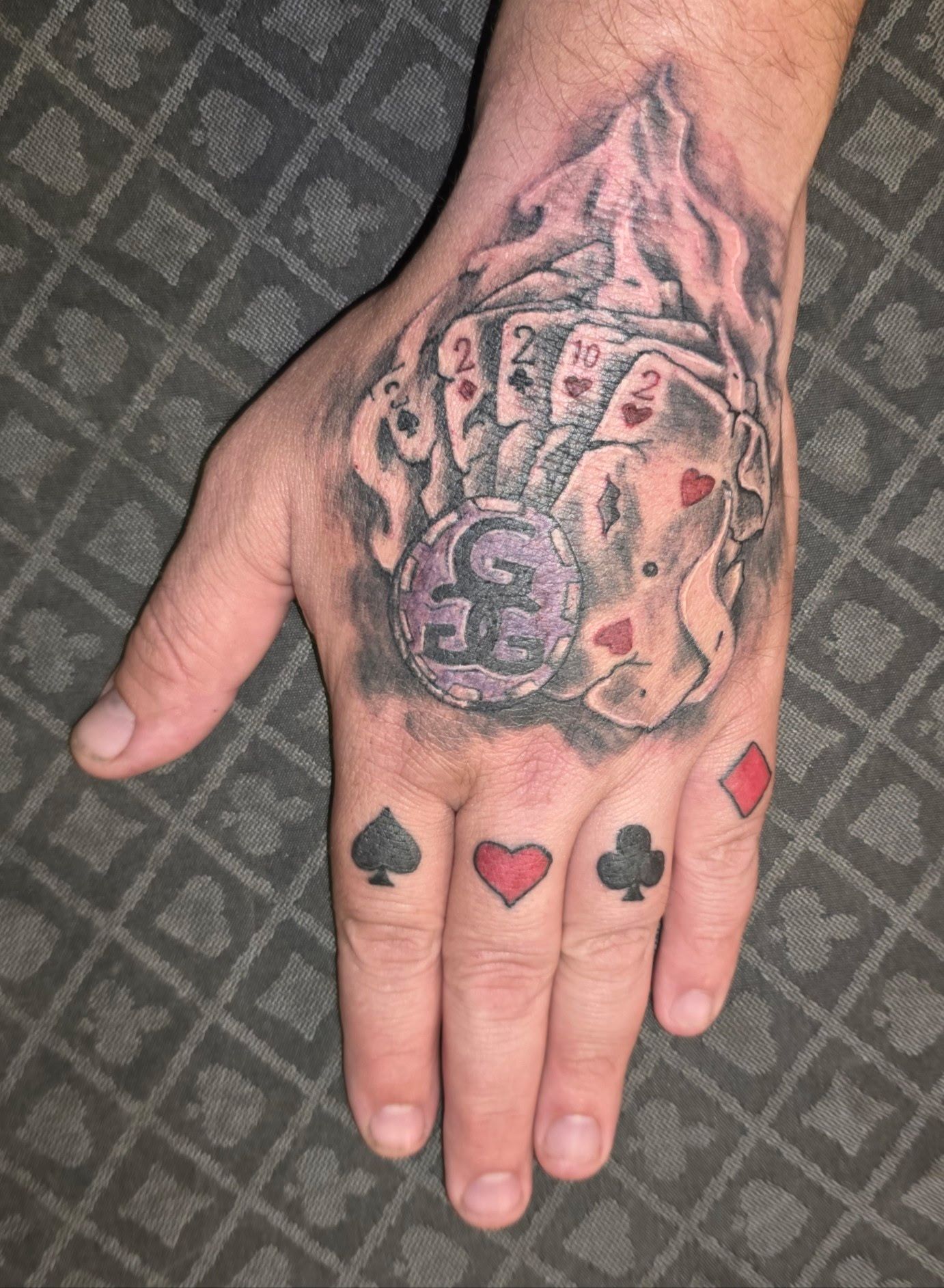 Hand with a dark tattoo on the back and playing card suit tattoos across the fingers.